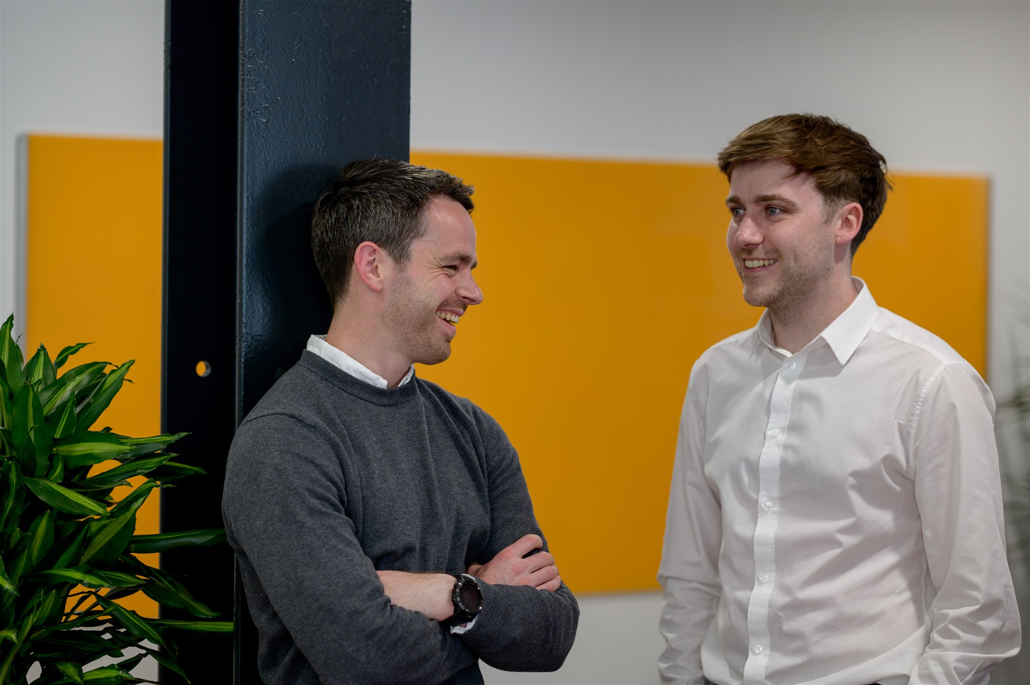 Two men in an office setting with an orange background wall smile at each other while speaking